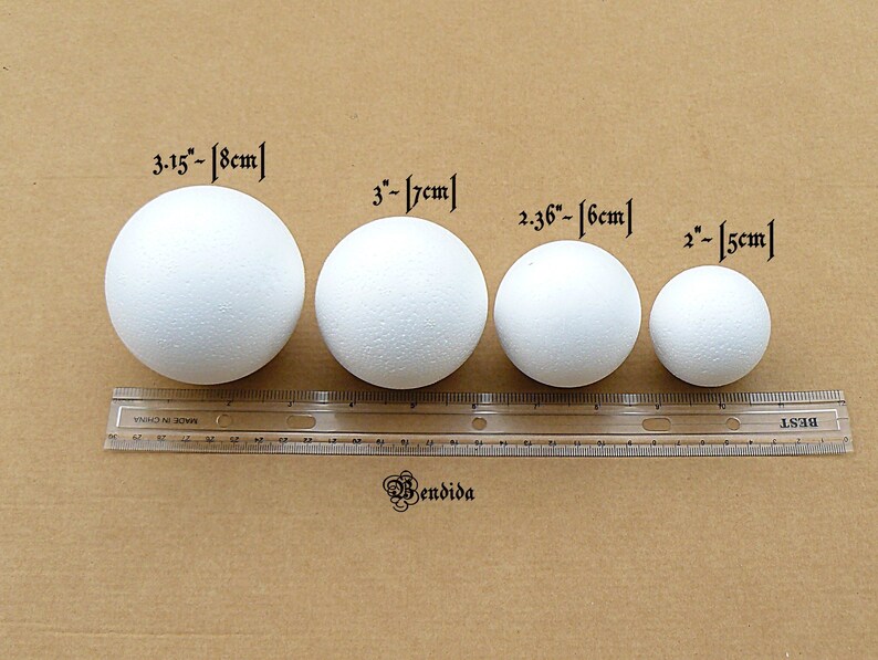 May include: Four white styrofoam balls of different sizes, measured in inches and centimeters, on a brown surface with a ruler.