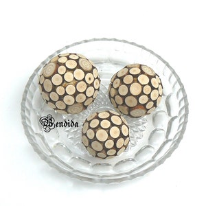 May include: Three decorative wooden spheres made of small, round pieces of wood arranged in a circular pattern. The spheres are displayed on a clear glass plate with a scalloped edge.