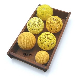 May include: A wooden tray containing six yellow and orange yarn balls. The balls are made from woven yarn and are arranged in a circular pattern. The tray is brown and has a rustic appearance.