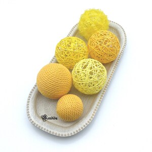 May include: A white oval tray with a beaded edge holds five yellow and cream-coloured yarn balls. The yarn balls are arranged in a semi-circle with a fluffy yellow pom-pom at the top of the tray.