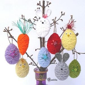 Puede incluir: Adornos de Pascua, incluyendo un conejo blanco, una zanahoria naranja y varios huevos envueltos en hilo de color morado, rojo, azul, amarillo y verde. Los adornos están suspendidos de ramas, creando una exhibición festiva. Se ve un jarrón morado con diseños.