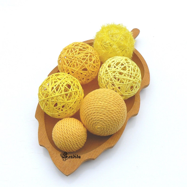 May include: A wooden bowl shaped like a leaf holds six yellow and light yellow balls of yarn. The balls are arranged in a cluster in the centre of the bowl.