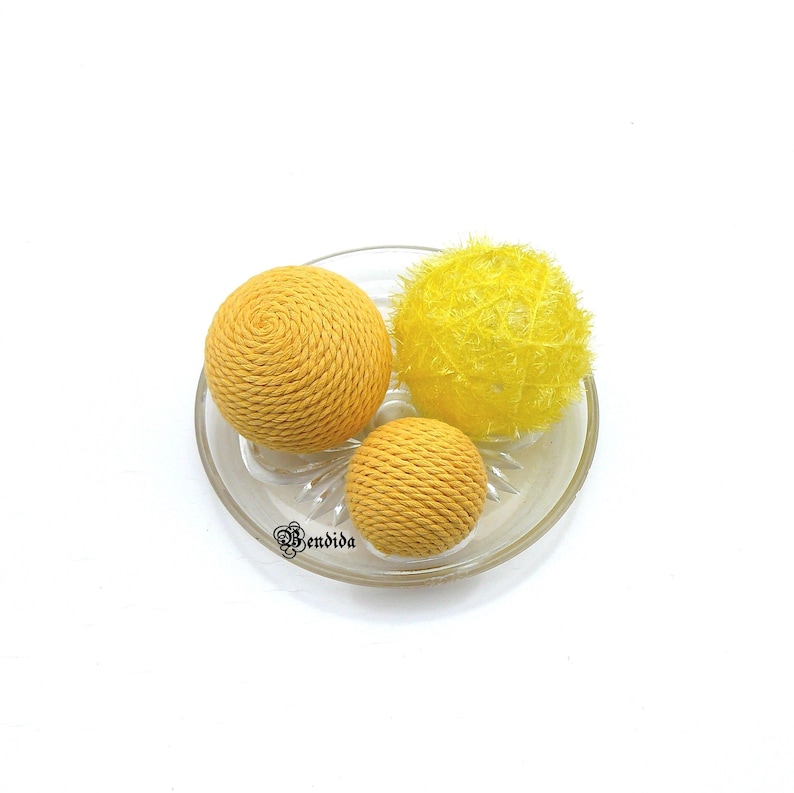 May include: Three yellow decorative balls on a clear glass plate. Two of the balls are made of woven rope and one is made of fluffy yarn.