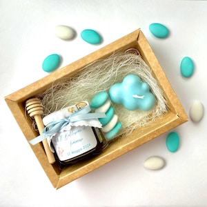 Può includere: Una scatola di cartone marrone piena di caramelle blu e bianche, un piccolo barattolo di miele con un mestolo di legno per il miele, una candela blu a forma di nuvola e carta bianca sminuzzata. Il barattolo ha un'etichetta che recita "Il Battesimo di Tommaso 12 Maggio 2024".