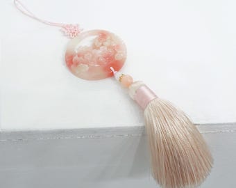 May include: A pink and white floral pendant with a tassel. The round pendant features carved flowers and is attached to a pink tassel with a matching cord and knot. The tassel is made of a soft, silky material.