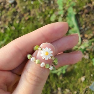 May include: Two beaded rings. One ring features a white daisy flower with a yellow center. The other ring has green beads and small white pearls. The rings are held up against a blurred green background.