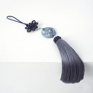 Könnte beinhalten: Ein dekorativer Quastenanhänger mit blau-grauem Farbverlauf, befestigt an einem ovalen Anhänger mit floralem Design. Die Quaste ist mit einem dunkelblauen Knoten und einer Schlaufe verbunden.