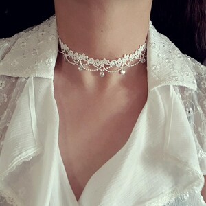 May include: A white lace choker necklace featuring a delicate floral pattern along the top edge, accented with thin dangling chains and small clear circular crystals spaced evenly along the bottom edge of the lace band.