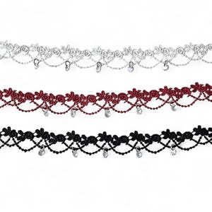 May include: Three strips of decorative lace trim in silver, dark red, and black. Each strip features a floral rose pattern along the top edge, a scalloped lattice design below, and small, clear, circular crystal-like charms hanging from the bottom.
