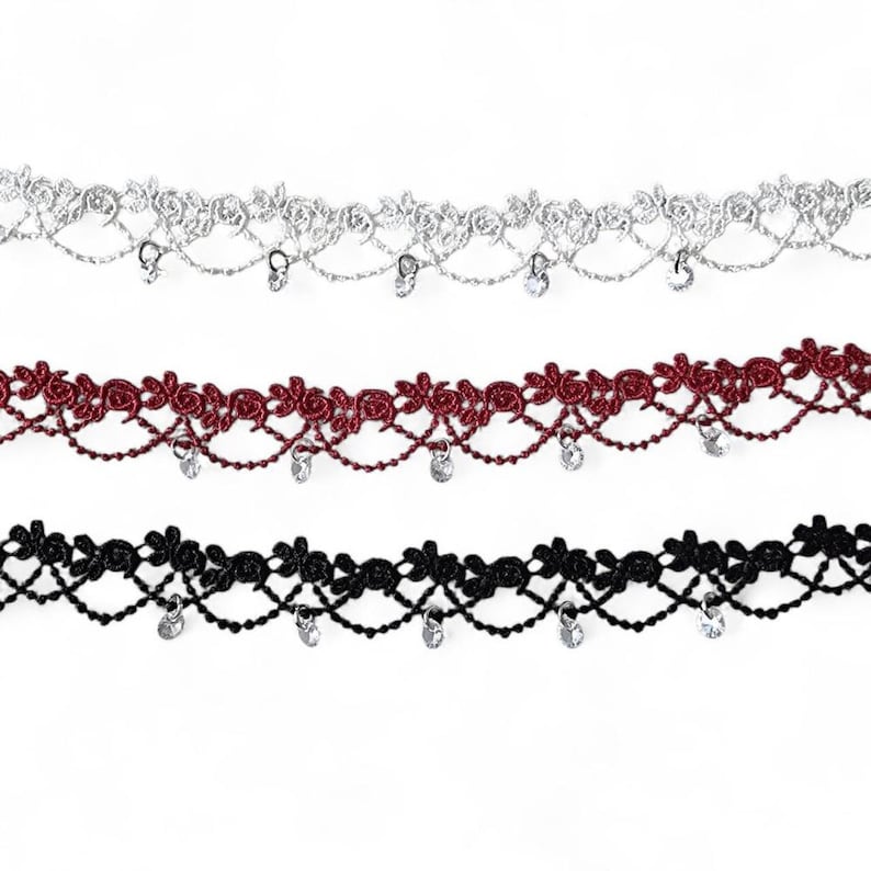 May include: Three strips of decorative lace trim in silver, dark red, and black. Each strip features a floral rose pattern along the top edge, a scalloped lattice design below, and small, clear, circular crystal-like charms hanging from the bottom.