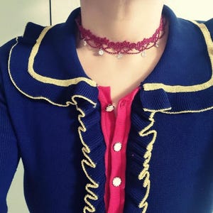 May include: A dark blue cardigan with gold trim, a pink button placket, and a red lace choker necklace featuring small dangling clear crystals. The cardigan has a ruffled collar and front edges, with three visible white buttons down the center.
