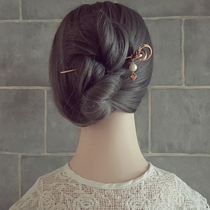 May include: A bun hairstyle secured with two decorative hairpins. One hairpin is a simple gold stick, and the other is a gold hairpin with a circular design, a pearl, and a heart charm. The hair is dark gray, and the person is wearing a white lace top.