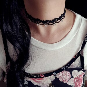 May include: A black lace choker necklace featuring a delicate floral pattern and small, clear, dangling crystal-like beads spaced evenly along the bottom edge. The intricate lace design sits snugly against the neck, highlighting the contrast between the dark fabric and the reflective, light-catching accents.