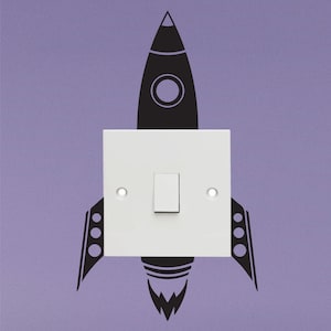 May include: A white light switch with a black rocket ship decal on a purple wall. The rocket ship has a black flame coming out of the bottom.