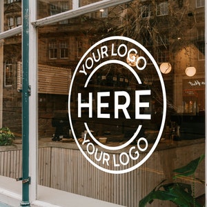 Op de afbeelding: Een etalage met een wit cirkelvormig logo-ontwerp met de tekst "YOUR LOGO HERE YOUR LOGO". De ruit weerspiegelt het interieur van een bedrijf, met een houten wand en planten.