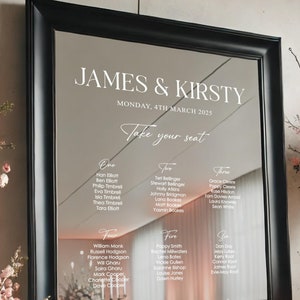 May include: A black framed mirror with a white inscription that reads "JAMES & KIRSTY, MONDAY, 4TH MARCH 2025, Take your seat". The mirror is reflecting a seating chart with names listed under table numbers one through eight.