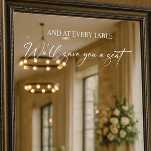 May include: A large, rectangular mirror with a dark, ornate frame. The mirror reflects a room with tables set for a formal event. White script on the mirror reads, "AND AT EVERY TABLE We'll save you a seat."