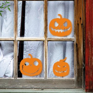 May include: Three orange jack-o'-lantern cutouts are displayed in a window. The cutouts are different sizes and have different facial expressions.