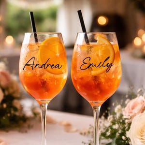 May include: Two stemmed glasses filled with an orange-colored beverage, garnished with orange slices and black straws. The name "Andrea" is written on one glass, and "Emily" on the other. The glasses sit on a white tablecloth, surrounded by flowers.