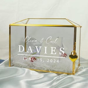 May include: A rectangular glass card box with a gold metal frame and a small gold heart-shaped padlock. The front glass panel features white printed text that reads Olivia & Carl DAVIES JUNE 15, 2024. The box contains white paper filler.
