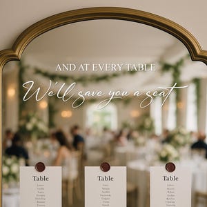 May include: A gold-framed mirror with the text "AND AT EVERY TABLE, We'll save you a seat." Six cards with guest names are attached. The background shows a blurred wedding reception.