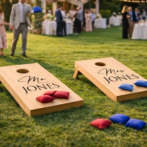 May include: Two wooden cornhole boards with the text "Mr JONES" and "Mrs JONES" in black script. Red and blue bean bags are placed on the boards and on the grass. The boards are set up on a green lawn, with people and a marquee in the background.