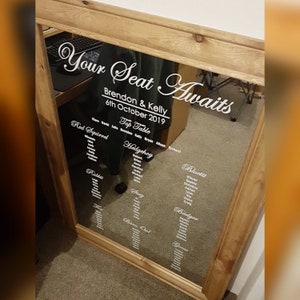 Puede incluir: Un espejo con marco de madera con un plano de asientos para una boda. El espejo tiene una pegatina de vinilo blanco que dice "Your Seat Awaits" con los nombres Brendon & Kelly y la fecha 6 de octubre de 2019. El plano de asientos enumera mesas con nombres como Red Squirrel, Hedgehog, Bluetit y Badger.