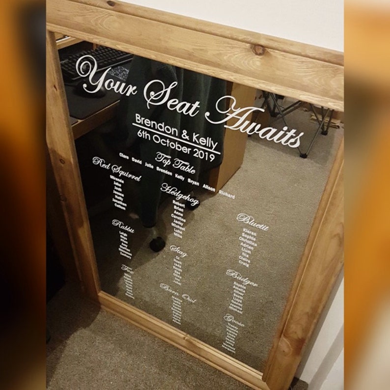 Puede incluir: Un espejo con marco de madera con un plano de asientos para una boda. El espejo tiene una pegatina de vinilo blanco que dice "Your Seat Awaits" con los nombres Brendon & Kelly y la fecha 6 de octubre de 2019. El plano de asientos enumera mesas con nombres como Red Squirrel, Hedgehog, Bluetit y Badger.