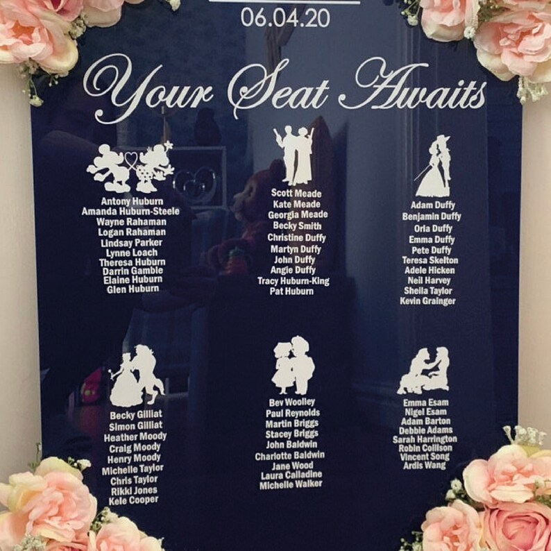 Puede incluir: Un letrero de acr&iacute;lico negro con texto e ilustraciones blancas. El letrero dice "Your Seat Awaits" y tiene un plano de asientos con nombres listados debajo de diferentes personajes de Disney. La fecha 06.04.20 est&aacute; en la parte superior del letrero.
