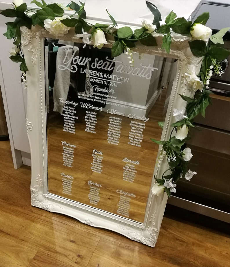May include: A white framed mirror with a seating chart for a wedding. The mirror has a white floral garland around it. The seating chart is written in white lettering on the mirror. The text on the mirror reads "Your seat awaits Lauren & Matthew March 31, 2018 Hendrick's Tanqueray W Edwards Bloems Bombay  Coerunn Oplh Sipsmith Puister Gordon's Beefeater"