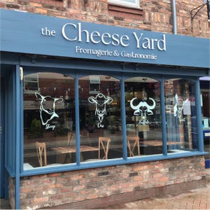 Może przedstawiać: Fasada sklepu "the Cheese Yard" z niebieską markizą i ceglaną fasadą. Nazwa sklepu jest w białych literach. Okna prezentują ilustracje owcy, krowy, bawołu i kozy.