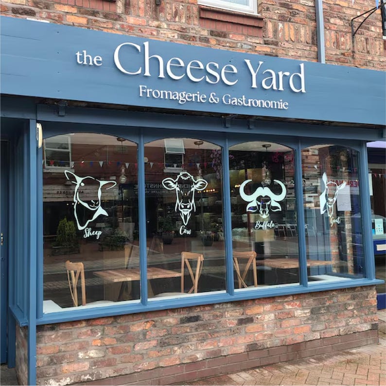 Może przedstawiać: Fasada sklepu "the Cheese Yard" z niebieską markizą i ceglaną fasadą. Nazwa sklepu jest w białych literach. Okna prezentują ilustracje owcy, krowy, bawołu i kozy.