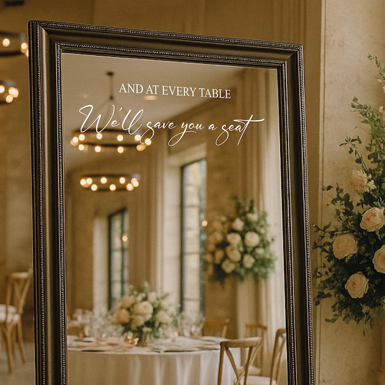 May include: A large, rectangular mirror with a dark, ornate frame. The mirror reflects a room with tables set for a formal event. White script on the mirror reads, "AND AT EVERY TABLE We'll save you a seat."
