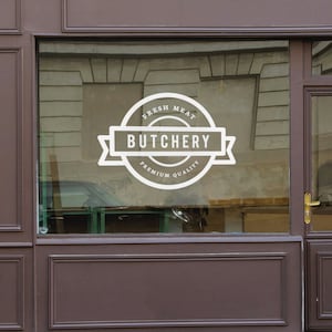 Może przedstawiać: Białe logo "Butchery" jest wyświetlane na szklanej witrynie sklepowej. Logo zawiera słowa "Fresh Meat" i "Premium Quality" w okrągłym kształcie. Witryna jest obramowana ciemnobrązowym drewnem.