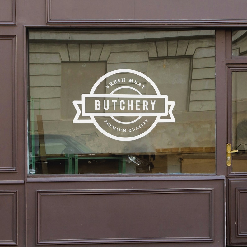 Może przedstawiać: Białe logo "Butchery" jest wyświetlane na szklanej witrynie sklepowej. Logo zawiera słowa "Fresh Meat" i "Premium Quality" w okrągłym kształcie. Witryna jest obramowana ciemnobrązowym drewnem.