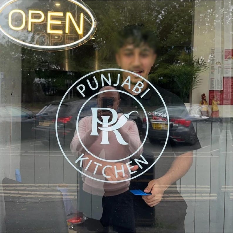 Może przedstawiać: Wystawa sklepowa z neonowym znakiem "OPEN". Okrągłe logo z napisem "PUNJABI KITCHEN" otacza stylizowany wz&oacute;r "PR". Odbicia ludzi i samochod&oacute;w są widoczne przez szybę.