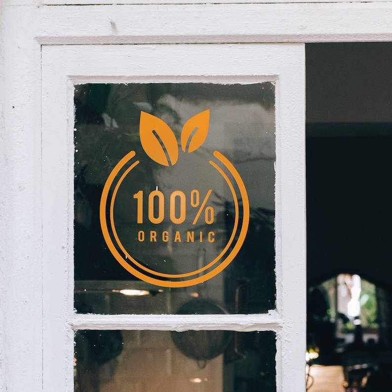 Może przedstawiać: Okno z białą ramą wyświetla pomarańczową naklejkę ze słowami "100% ORGANIC" wewnątrz okrągłego wzoru z dwoma liśćmi na g&oacute;rze. Naklejka jest przymocowana do szyby okiennej.