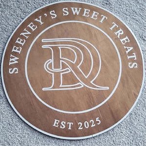 Może przedstawiać: Okrągły drewniany znak z napisem "Sweeney's Sweet Treats" w białym kolorze. Znak posiada stylizowany biały monogram "SR" na środku oraz napis "EST 2025" na dole. Znak ma fakturę słoj&oacute;w drewna i brązowy kolor.