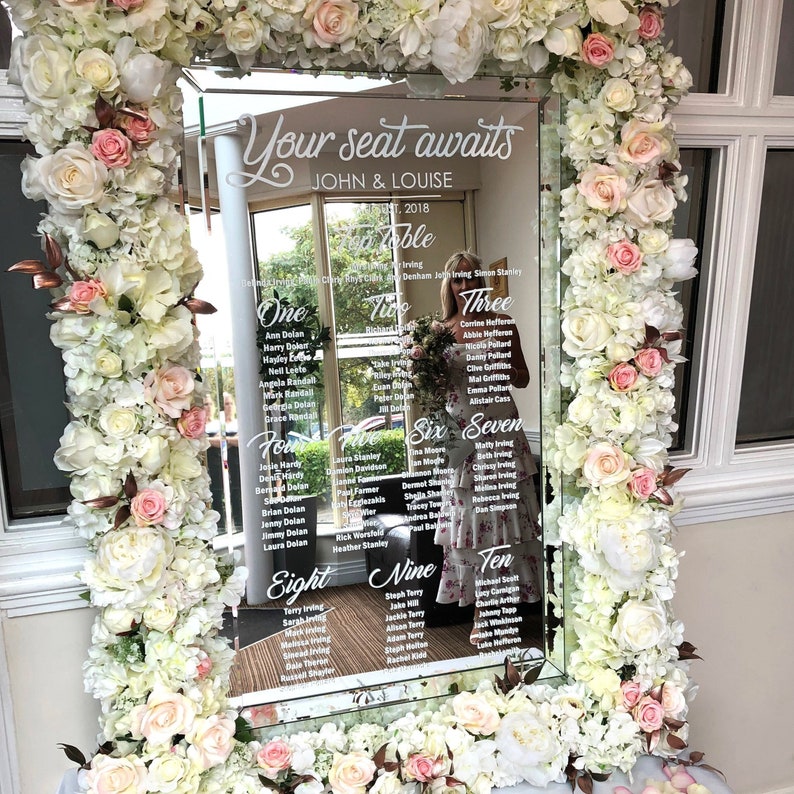 May include: A white mirror with a floral frame and a seating chart for a wedding reception. The chart is titled "Your seat awaits John & Louise" and lists the names of guests at each table.