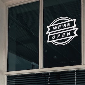 Może przedstawiać: Biały znak "We're Open" jest przymocowany do okna. Znak zawiera tekst w projekcie banera w okrągłej ramie. Okno jest podzielone na dwie szyby, z żaluzjami widocznymi na dole.