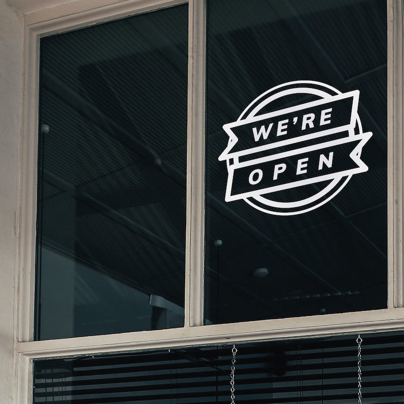 Może przedstawiać: Biały znak "We're Open" jest przymocowany do okna. Znak zawiera tekst w projekcie banera w okrągłej ramie. Okno jest podzielone na dwie szyby, z żaluzjami widocznymi na dole.