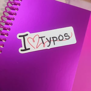 May include: A purple spiral-bound notebook with a sticker that reads "I ❤️ Typos." The sticker has black text and a red heart. The notebook is a vibrant shade of purple.
