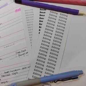 May include: A sheet of white rectangular stickers featuring the word Editing printed in a black typewriter-style font. The sticker sheet is surrounded by several pens in shades of pink, purple, and blue, along with a pink metal binder clip.
