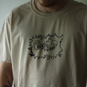 May include: A beige t-shirt with a black graphic print of two faces with the text "BLABLABLABLA" repeated around them.
