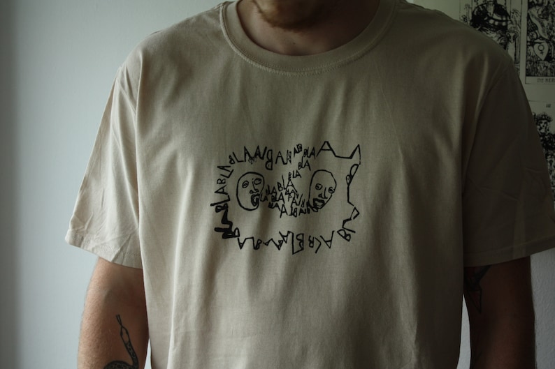 May include: A beige t-shirt with a black graphic print of two faces with the text "BLABLABLABLA" repeated around them.