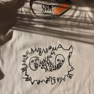 May include: A beige t-shirt with a black graphic print of two faces with the text "BLABLABLA" around them.