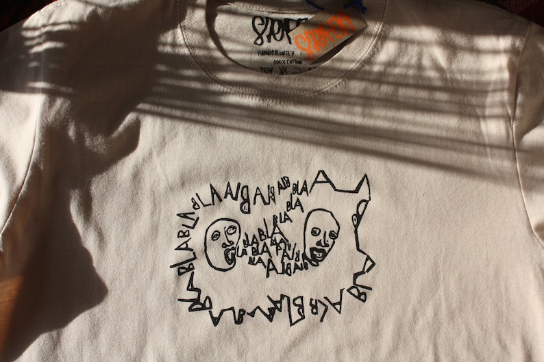 May include: A beige t-shirt with a black graphic print of two faces with the text "BLABLABLA" around them.