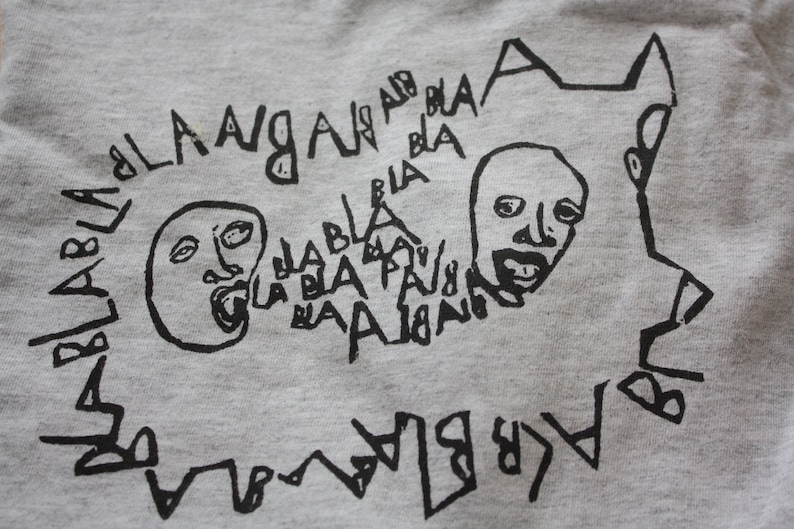 May include: A gray t-shirt with a black graphic print of two faces with open mouths and the words "bla bla bla" and "la la la" repeated around the faces.