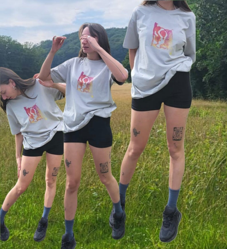 May include: Three people wearing light gray t-shirts with a colorful abstract design, paired with black shorts, blue socks, and black sneakers. The background is a grassy field with trees and a cloudy sky.