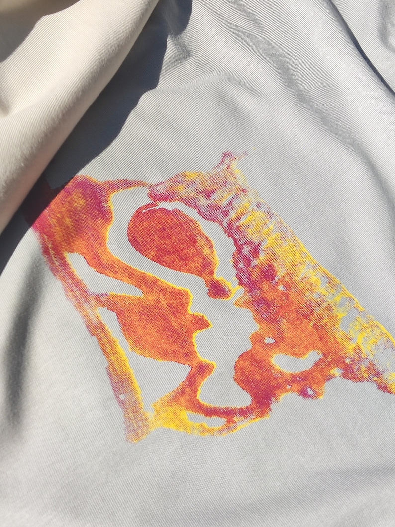 May include: Close-up of a light gray fabric with a vibrant abstract design in shades of orange, yellow, and pink. The design appears to be a stylized shape. A section of off-white fabric is visible in the upper left corner.